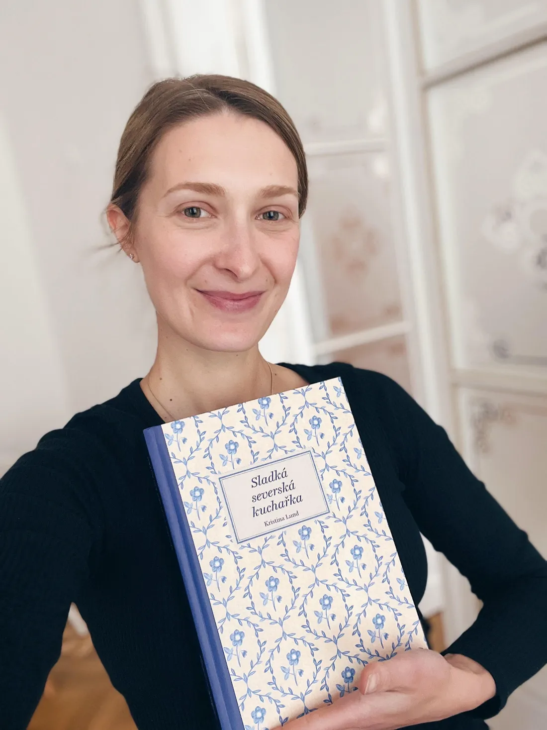 Kristina Lund se usmívá a drží kuchařku Sladká severská kuchařka s modrým květinovým vzorem