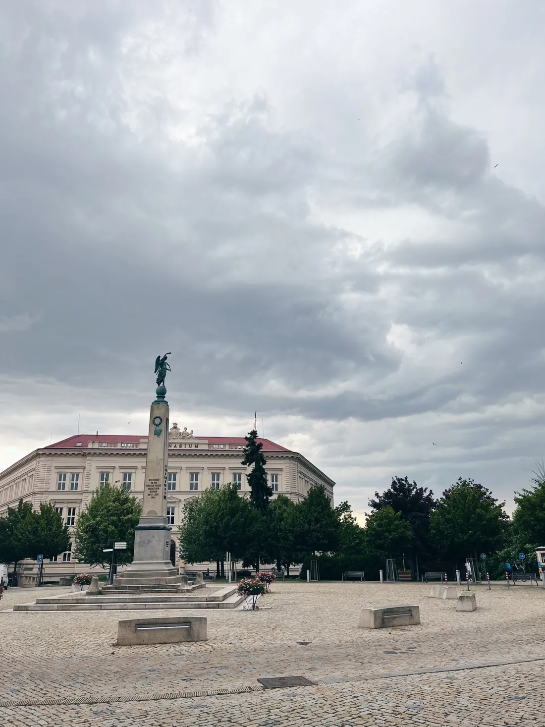 Komenského náměstí se sloupem a budovou gymnázia pod zataženou oblohou