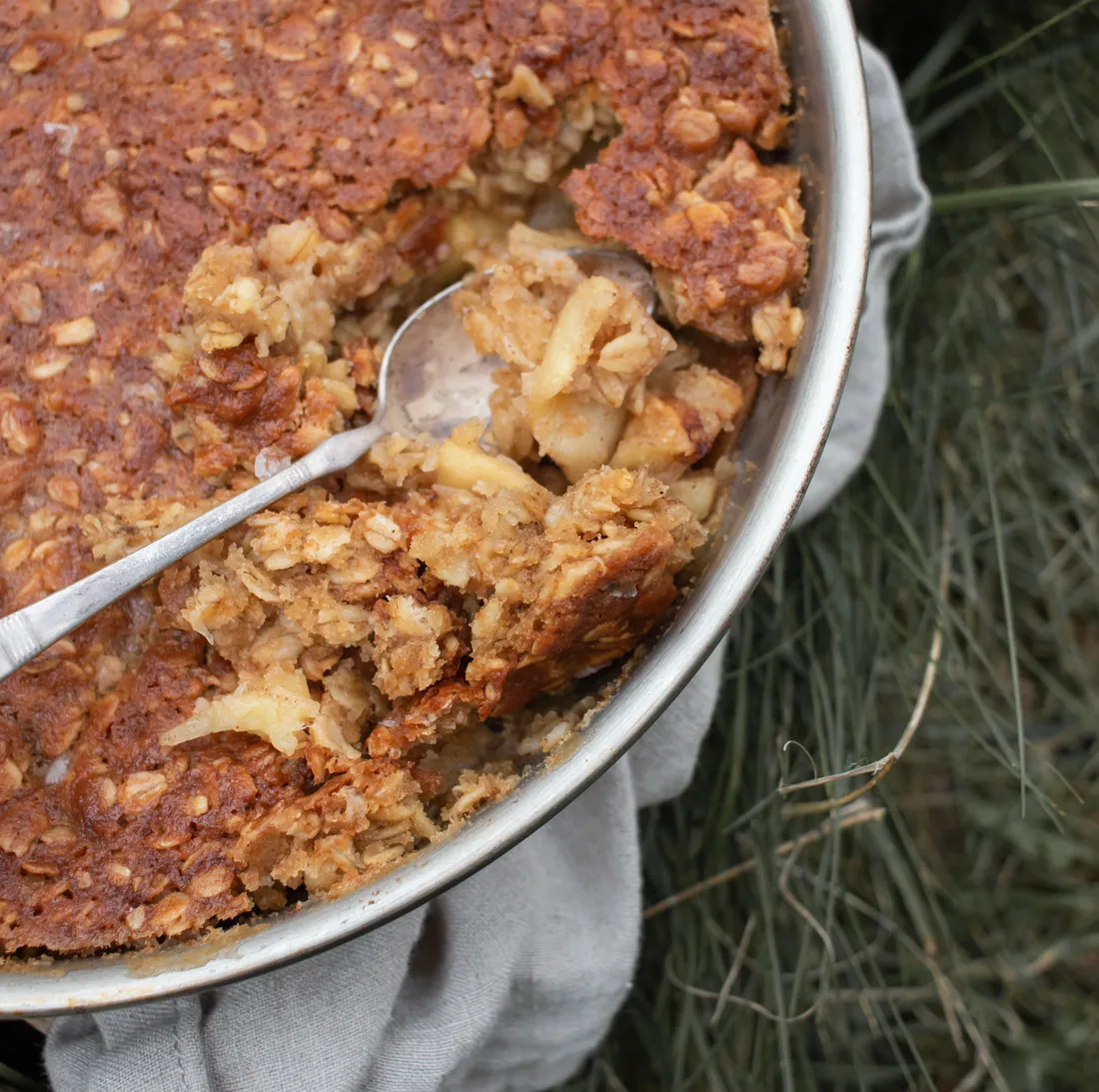 Zapečená ovesná kaše s jablky v kulaté formě na šedé utěrce v trávě, lžíce nabírá porci