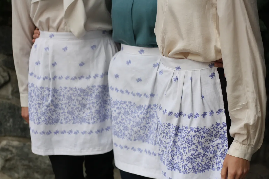 Tři ženy v bílých zástěrách s fialovým květinovým vzorem stojí objaté u kamenné zdi, detail pasů a rukou