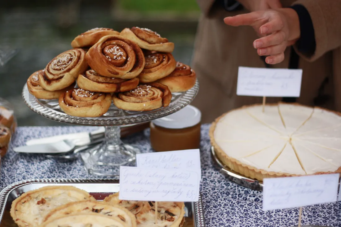 Skořicoví šneci na skleněném stojanu, krémový koláč a malé slané quiche s ručně psanými cenovkami na trhu
