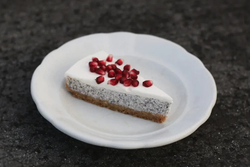 Řez makového cheesecaku s bílou vrstvou a zrnky granátového jablka na vlnitém talíři na tmavém kamenném podkladu
