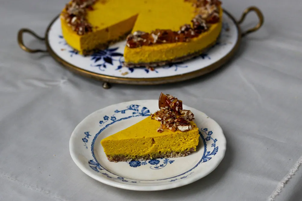 Kurkumový cheesecake s ořechy a karamelem na modrobílém talíři, celý dort a ukrojený kousek v popředí na ubrusu