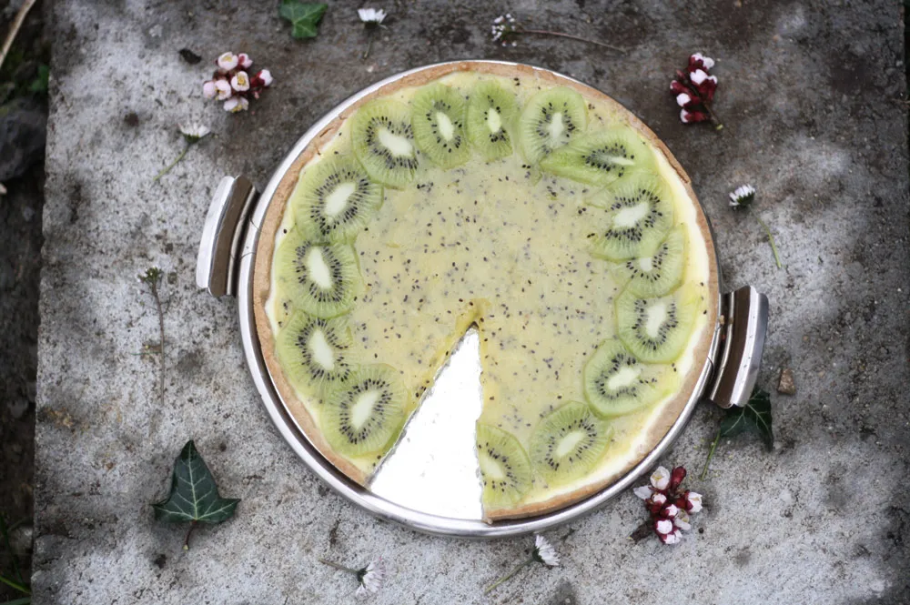 Kiwi koláč s plátky kiwi po obvodu a chybějícím kouskem na stříbrném podnosu mezi sedmikráskami a břečťanem