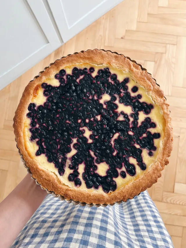 Kulatý borůvkový koláč s krémovou náplní a tmavými borůvkami držený v ruce nad parketami v modrém zástěře.
