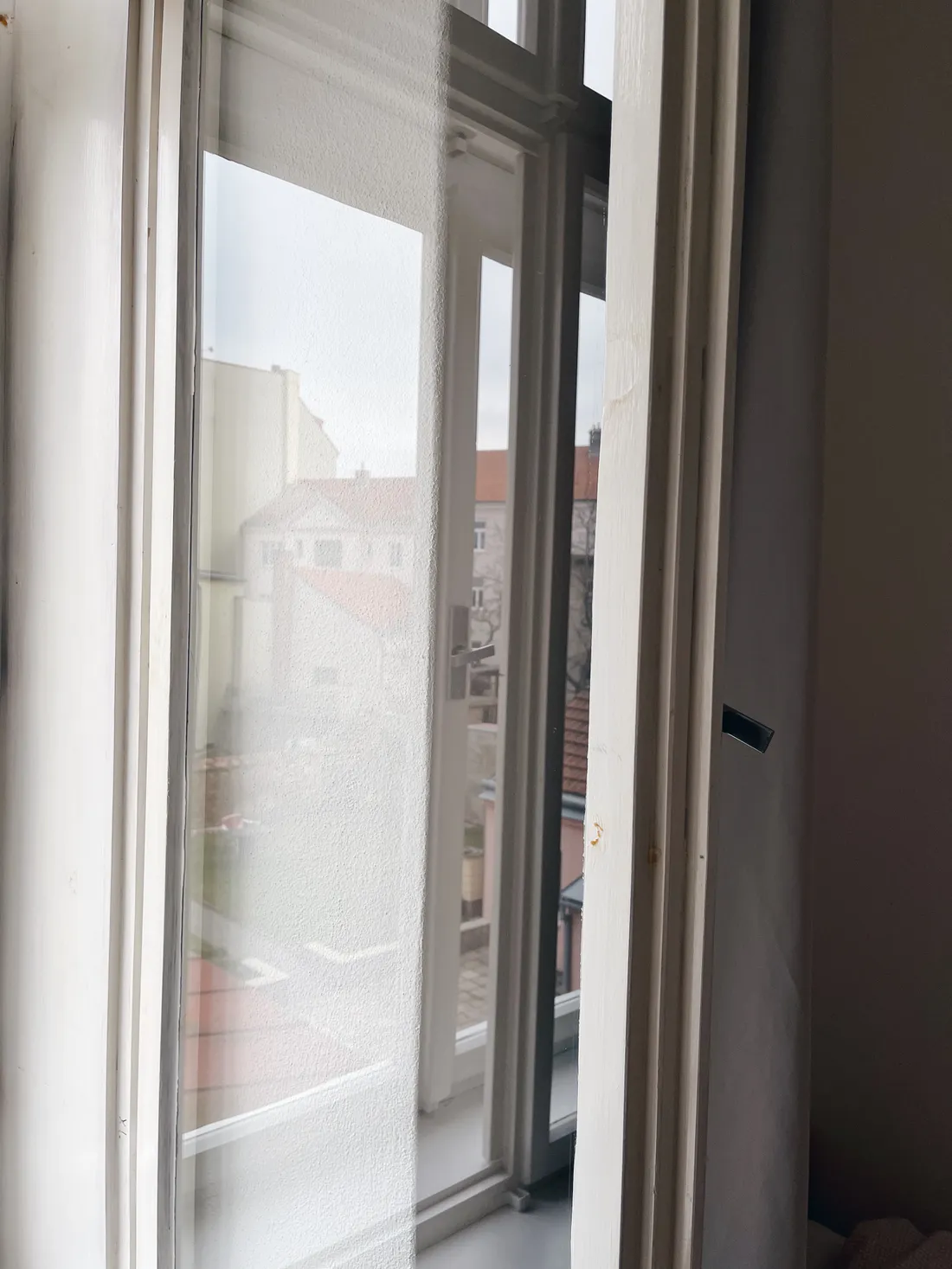 Otevřené bílé špaletové okno s výhledem na červené střechy a světlé fasády domů ve vnitrobloku.