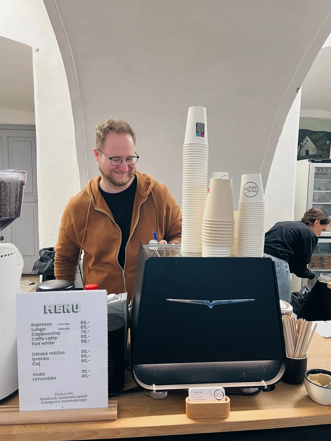 Barista za dubovým pultem u černého espresso přístroje a ceníku Káva na Knopp ve světnici.