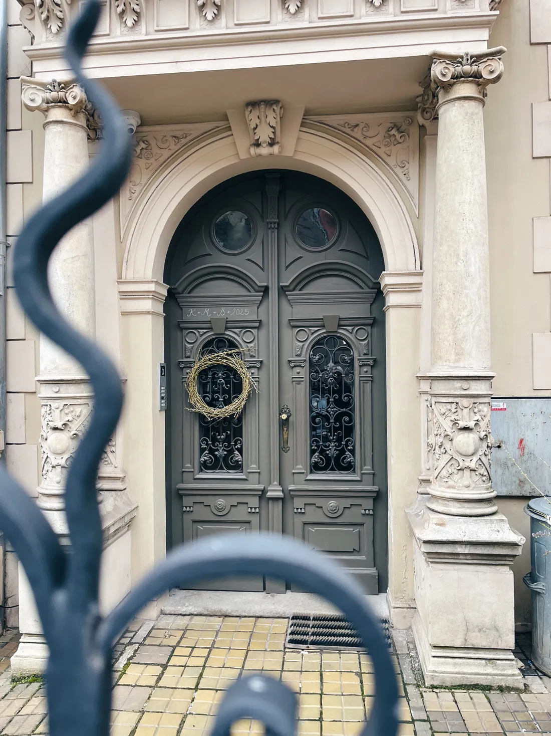 Zdobený kamenný portál s tmavými dveřmi, věncem a nápisem K+M+B nad kovovým plotem.