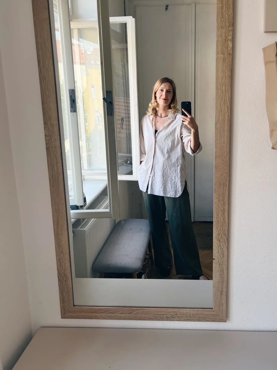 Zrcadlové selfie blonďaté ženy ve světlé pruhované košili a tmavě zelených širokých kalhotách v pokoji s oknem