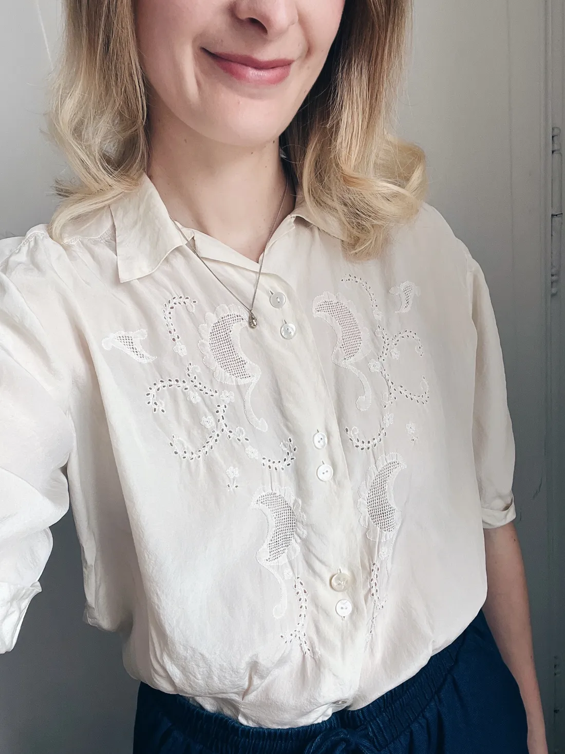 Detail krémové halenky s bílou výšivkou a dírkovaným vzorem, blond vlny a tenký perlový řetízek