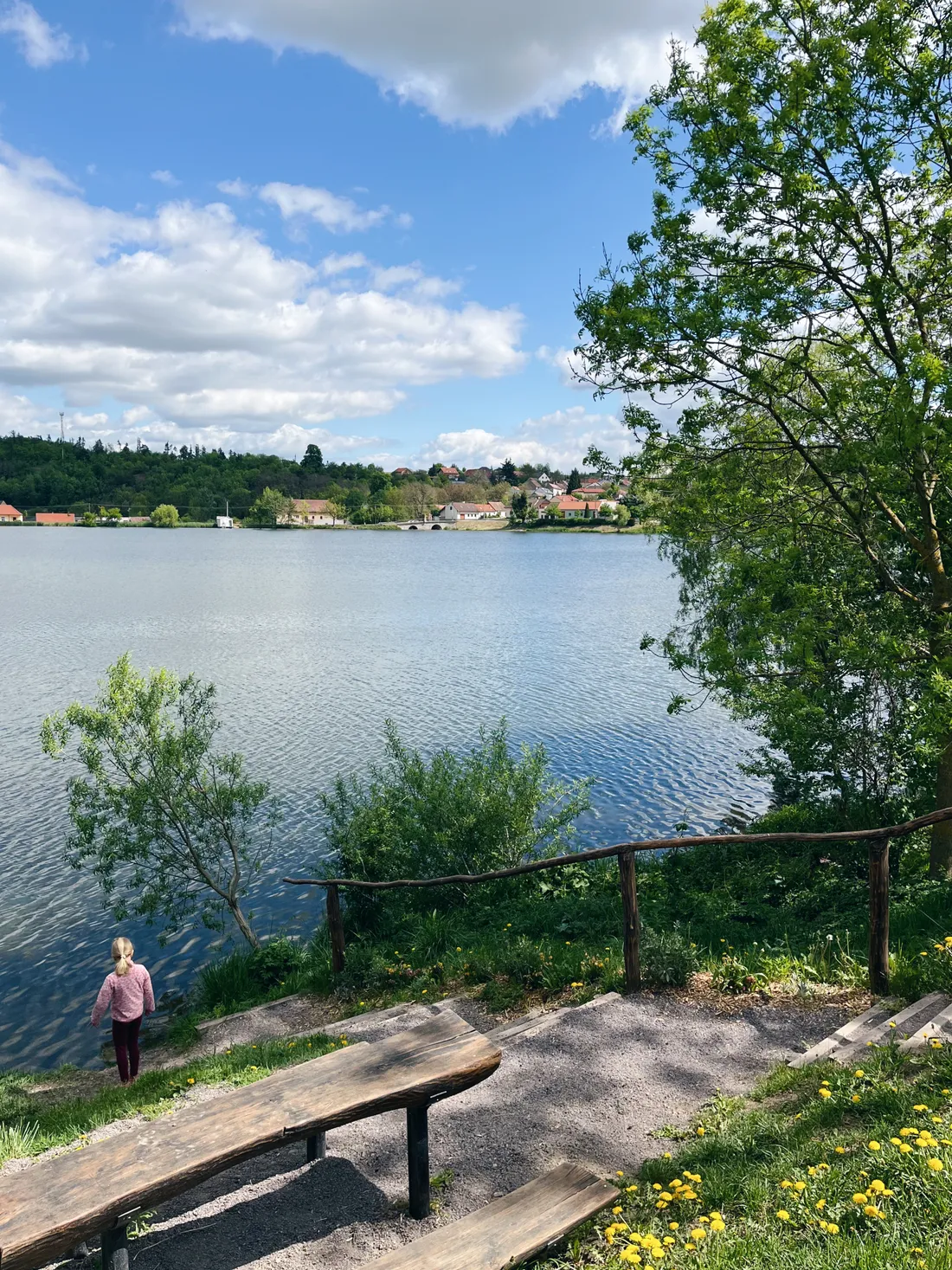 Jezero s vesnicí na protějším břehu, zelenými kopci a modrou oblohou, dřevěné lavičky v popředí