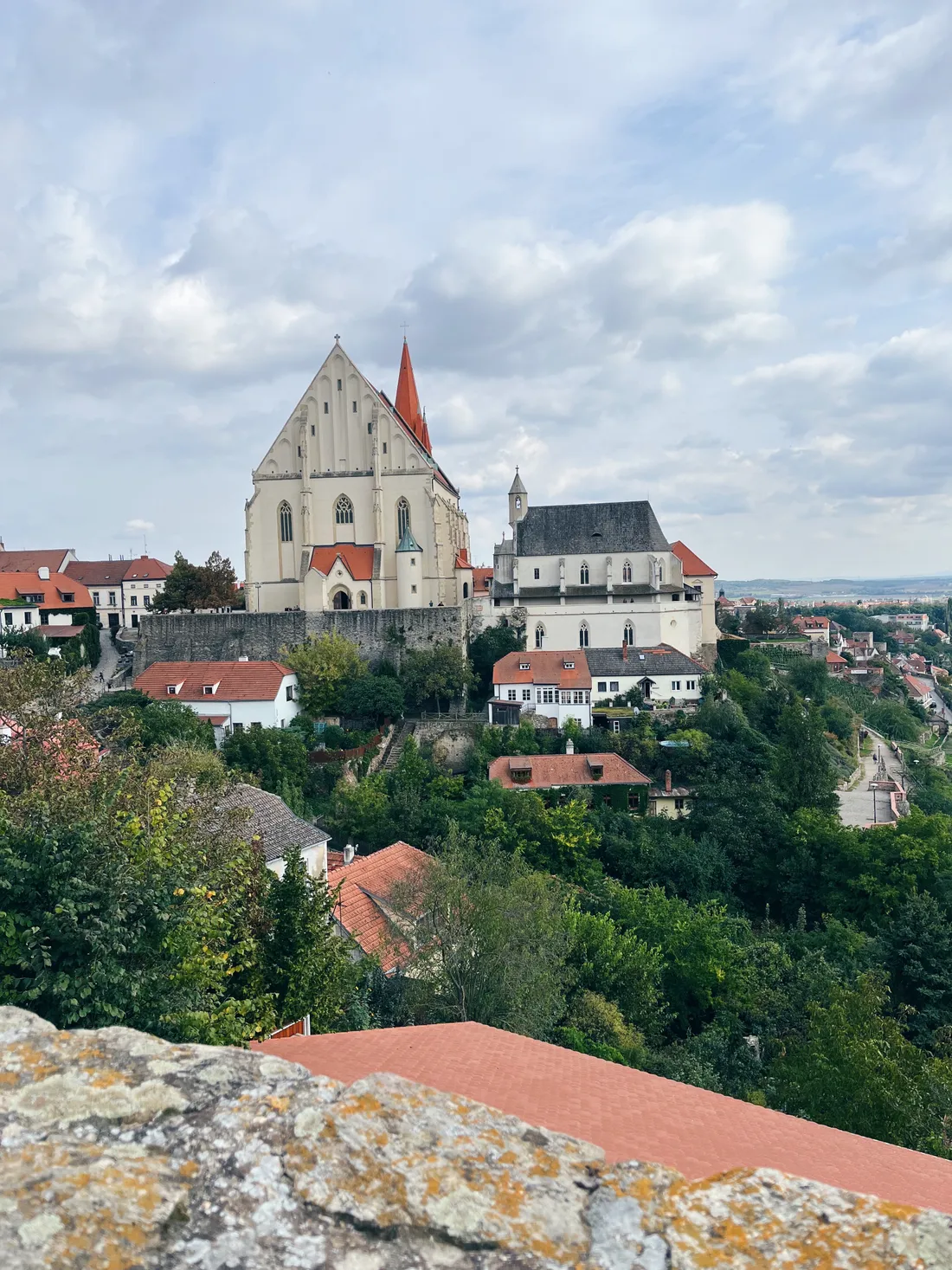 Kostel sv. Mikuláše s červenou věží na kopci nad bílými domy se střechami, pohled přes oblačnou krajinu.