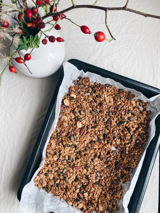 Zlatavá granola s pekany a dýňovými semínky na plechu; vedle váza se šípky.
