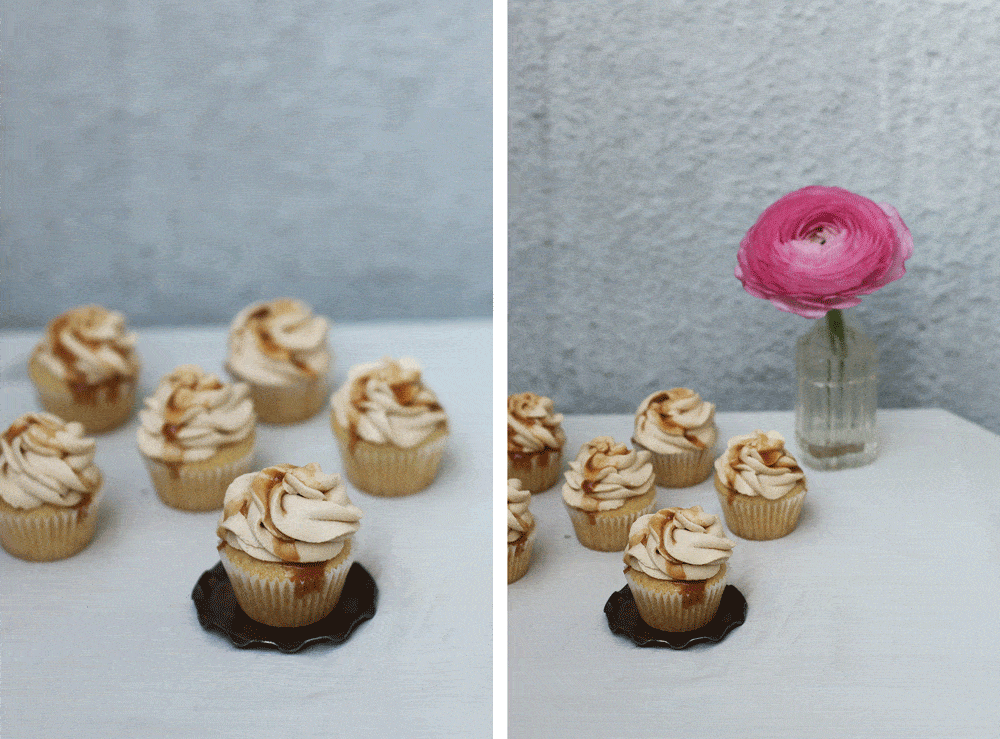 Karamelové cupcakes na bílém povrchu, jeden na tmavém talíři, vpravo růžový květ ve skleněné vázičce