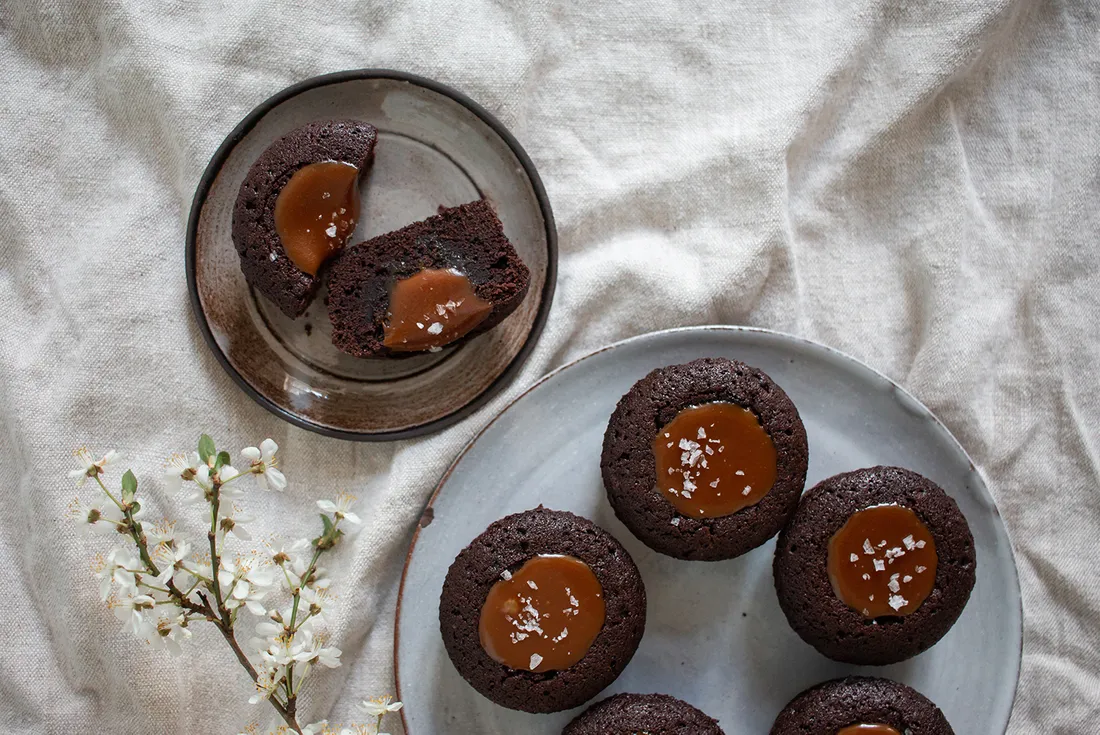 Pět muffinů s karamelem a solí na šedém talíři a rozkrojený muffin na hnědém talíři na lněném ubrusu
