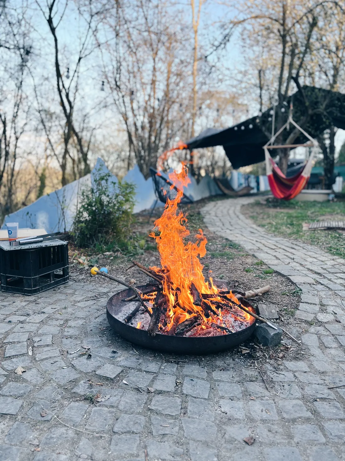 Plameny v kovovém ohništi na kamenné terase, v zahradě houpací síť a holé stromy.