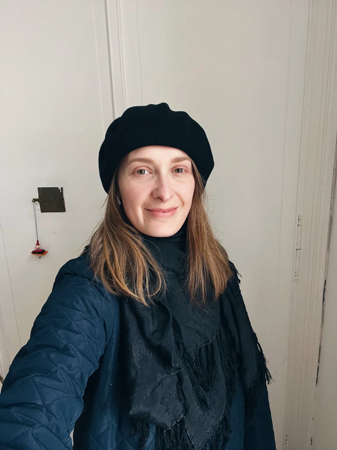Selfie usměvavé ženy v černém beretu, prošívané bundě a těžkém šále s brčkami.
