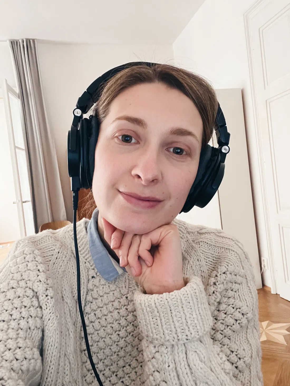 Žena v krémovém pleteném svetru s velkými černými sluchátky opírá bradu o ruku v bílém pokoji.