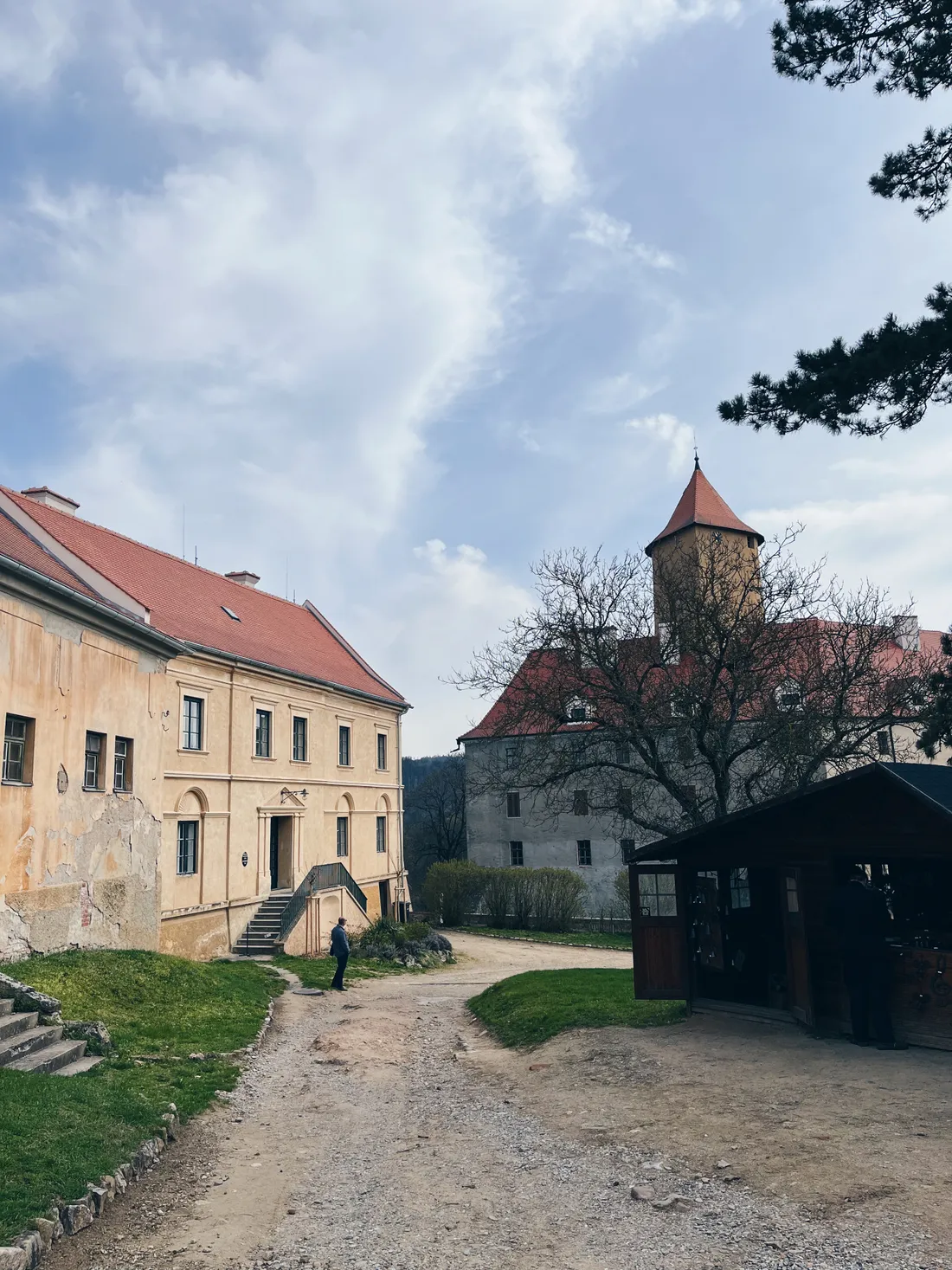 Nádvoří s žlutou budovou, šedým hradem s věží a prašnou cestou mezi trávníkem pod jasnou oblohou.