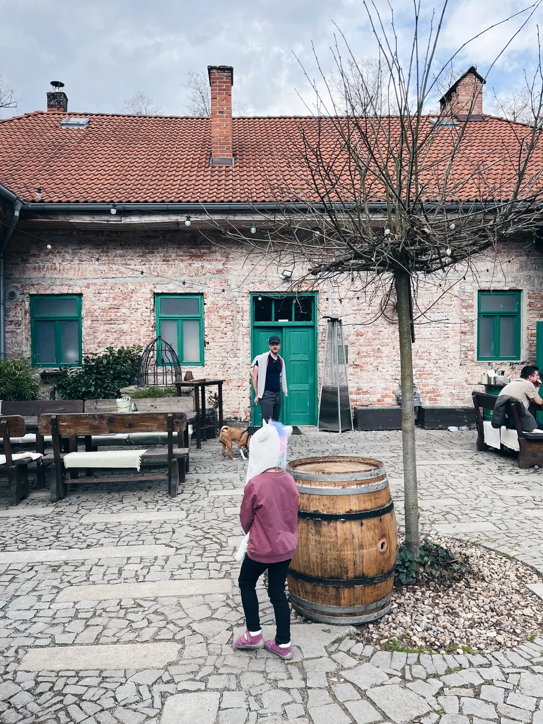 Dítě v růžové mikině s kapucí jednorožce na dlážděném dvoře před cihlovou budovou se zelenými okny.