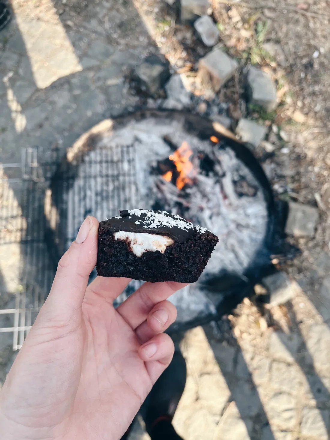 Ruka drží tmavý čokoládový cupcake s bílou náplní a kokosem u ohniště na zahradě.