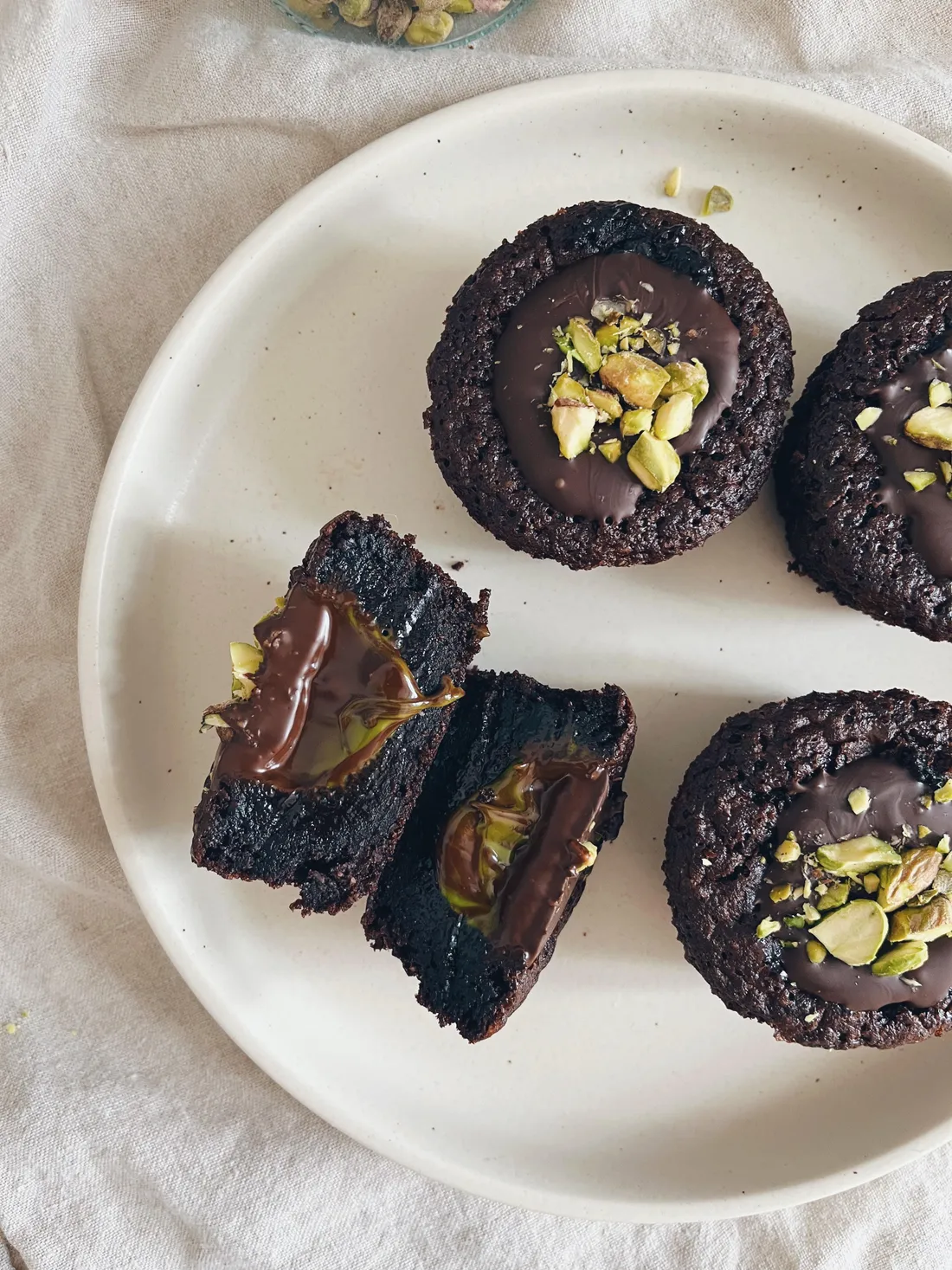 Čokoládové muffiny s tmavou ganache a sekanými pistaciemi na bílém talíři na lněném ubrusu.