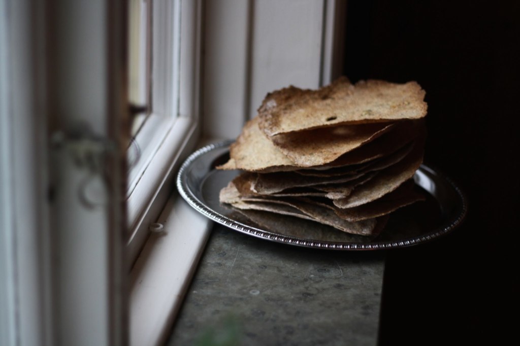 Stoh křupavých knäckebröd se sezamem na stříbrném podnosu u bílého okna se záclonou.