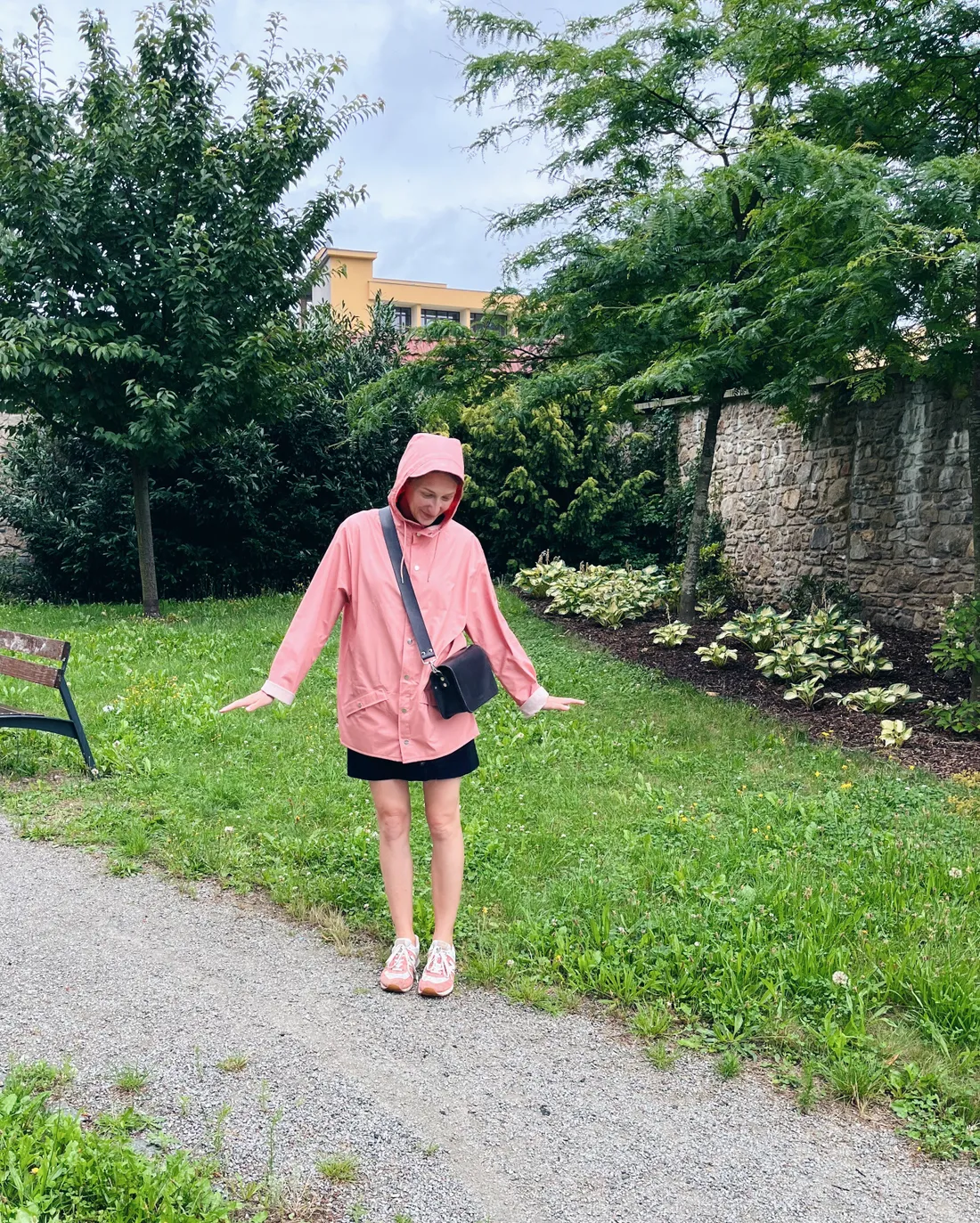 Žena v růžové pláštěnce s kapucí stojí na štěrkové cestě v parku se zelenou zahradou a stromy