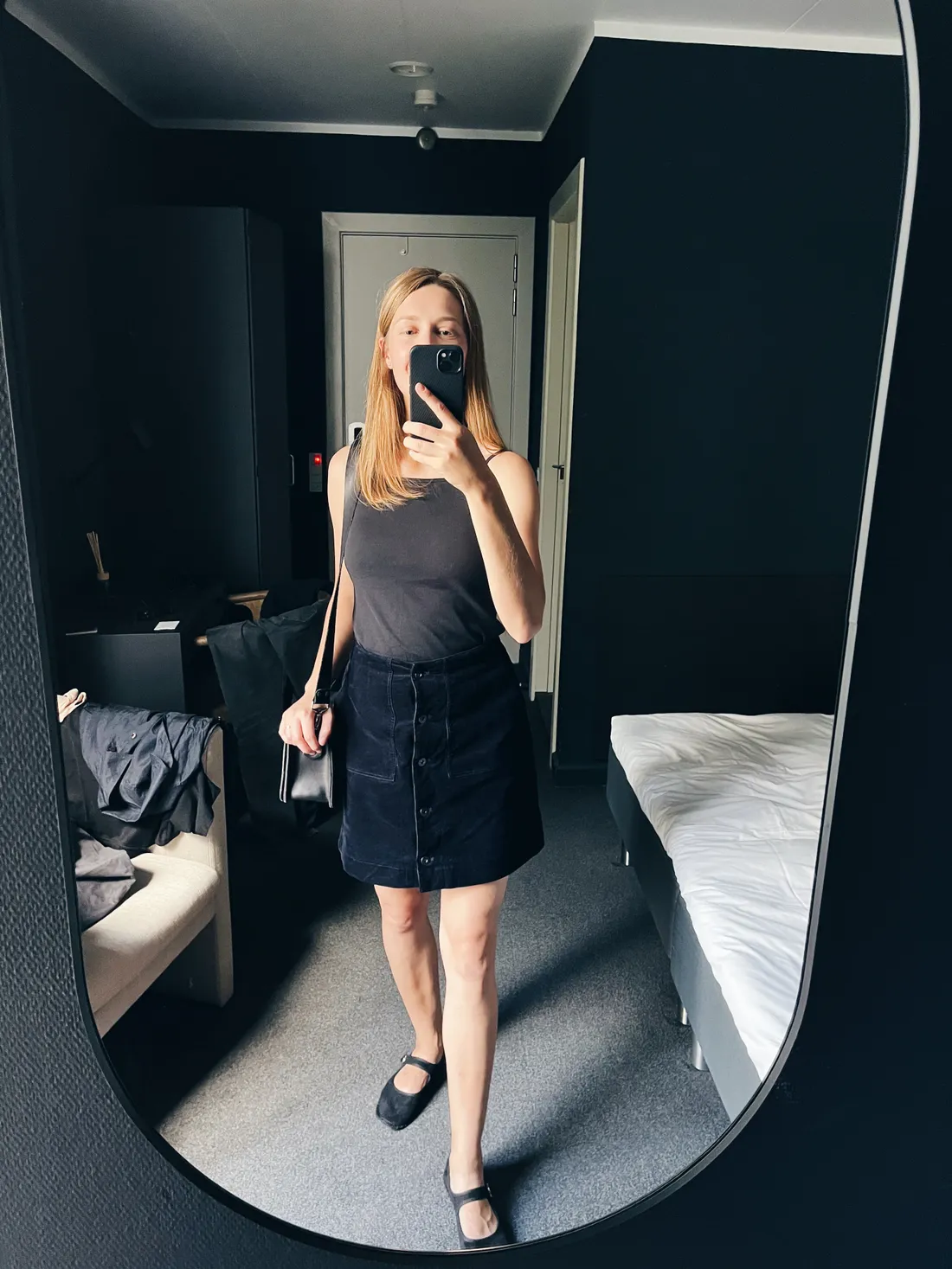 Blondýna v tmavém tílku a manžestrové suknici se fotí v zrcadle v tmavém hotelovém pokoji.