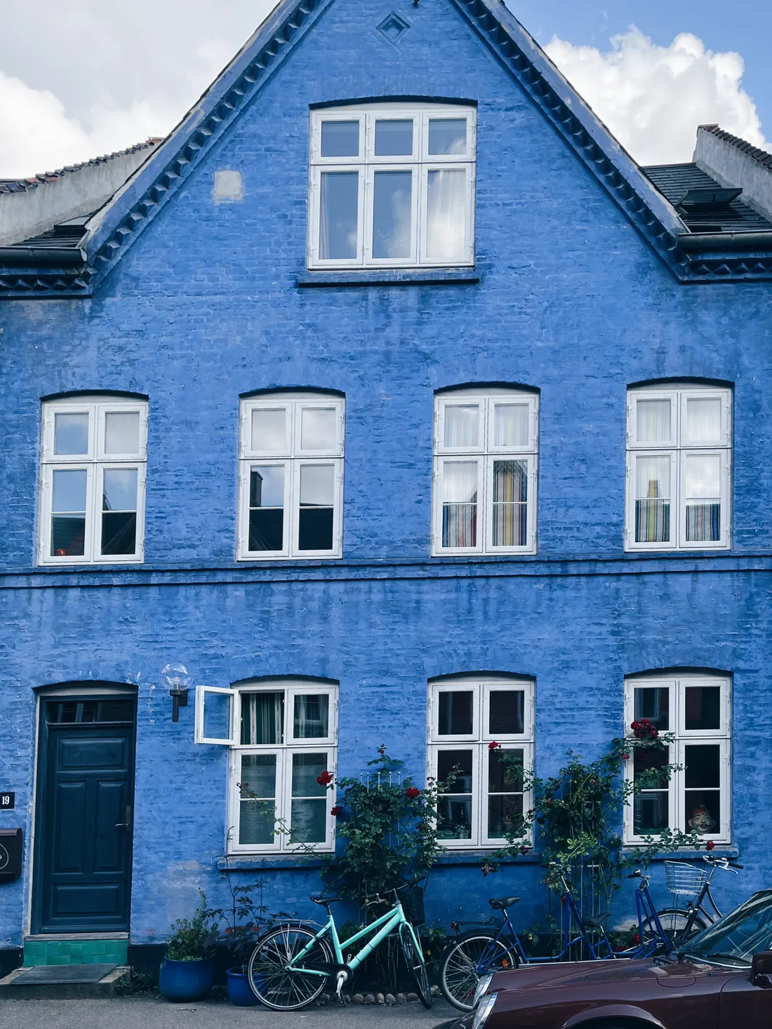 Jasně modrý cihlový dům s bílými okny, koly před vchodem a červenými růžemi u fasády.