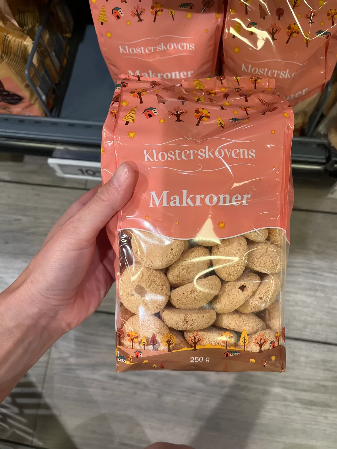 Ruka drží růžový sáček makronek Klosterskovens Makroner v obchodě s podzimním potiskem.