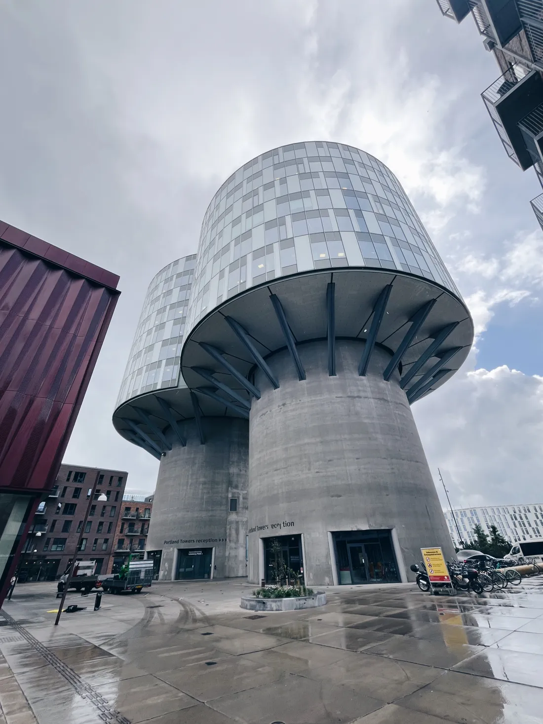 Dvě betonové Portland Towers s prosklenou nástavbou na mokré dlažbě pod zataženou oblohou.