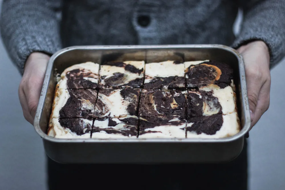 Pekáč mramorových cheesecake brownies v rukou osoby v šedém pleteném svetru.