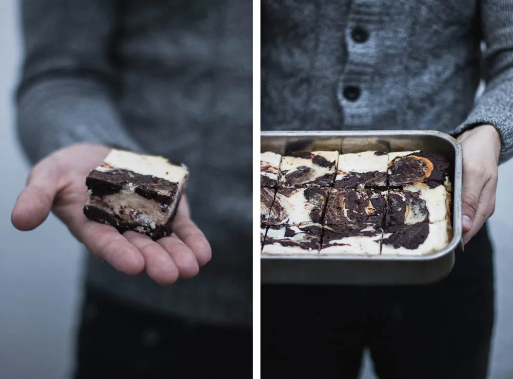 Kousek mramorového cheesecake brownie v dlani a celý nakrájený plech v rukou v šedém svetru.