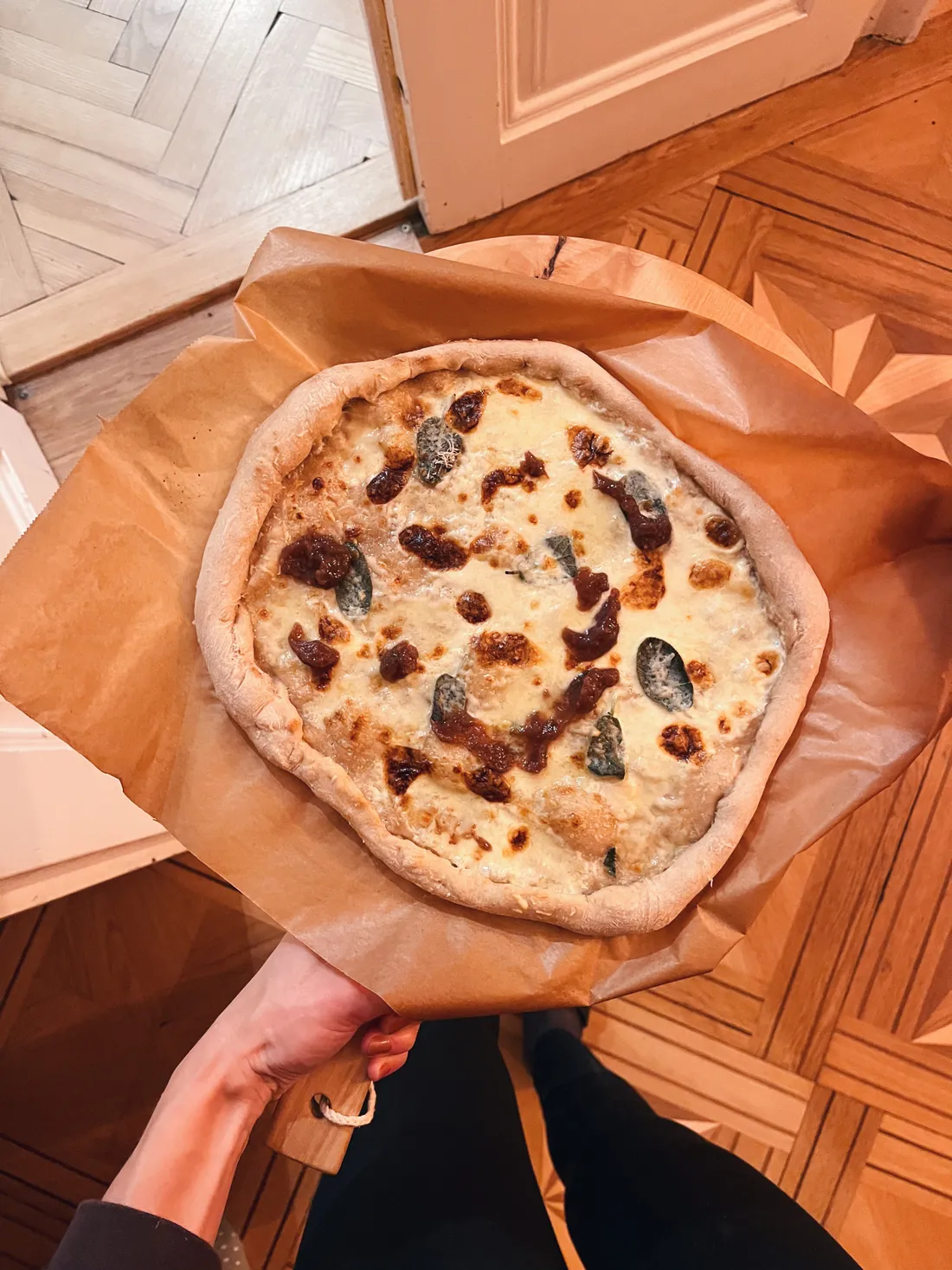 Domácí pizza se sýrem a tmavými skvrnami na pečicím papíru na dřevěné lopatě nad parketovou podlahou.