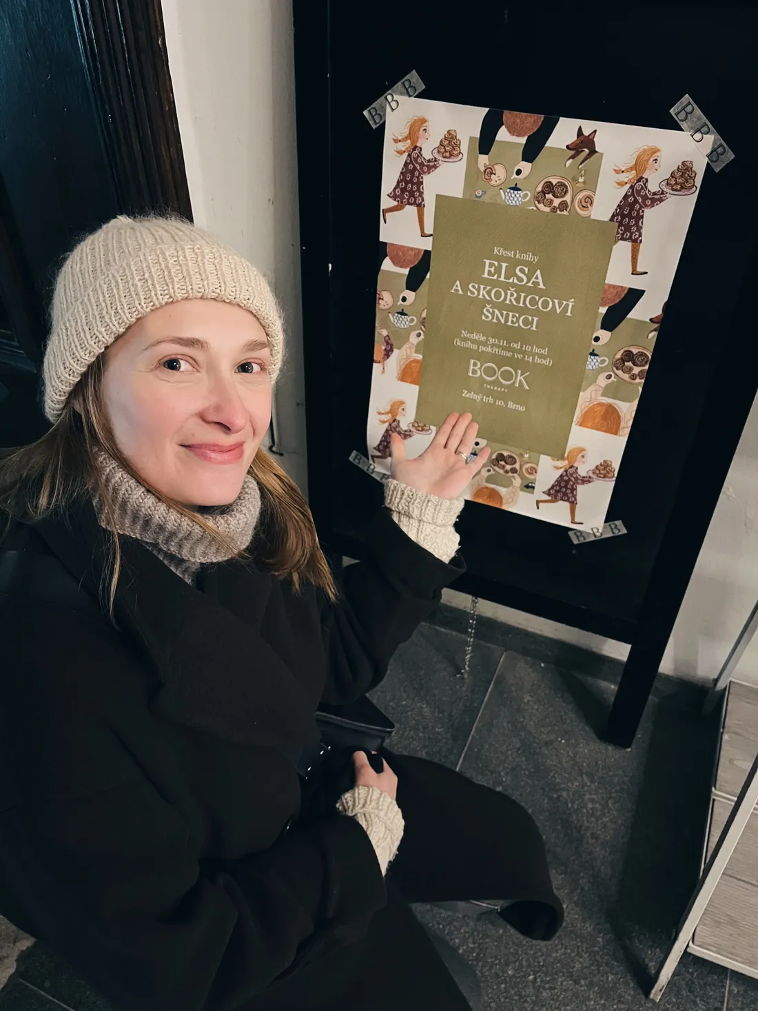 Žena v čepici ukazuje na plakát křtu knihy Elsa a skořicoví šneci v Book Therapy.