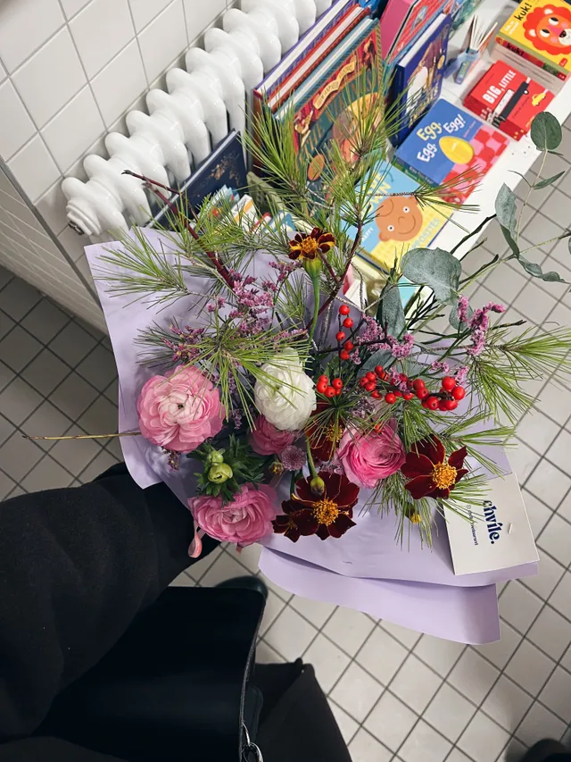 Kytice růžových a tmavých květů s borovicovým jehličím a bobulemi v lila papíru, držená v ruce u dětských knih.