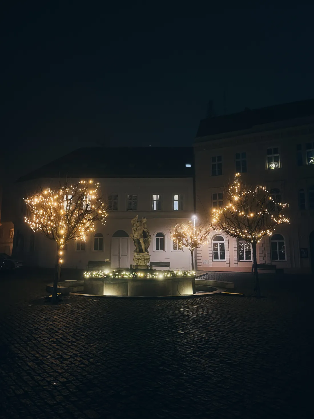 Náměstí v noci s osvětlenou kašnou, třemi stromy v světýlkách a barevnými fasádami domů.