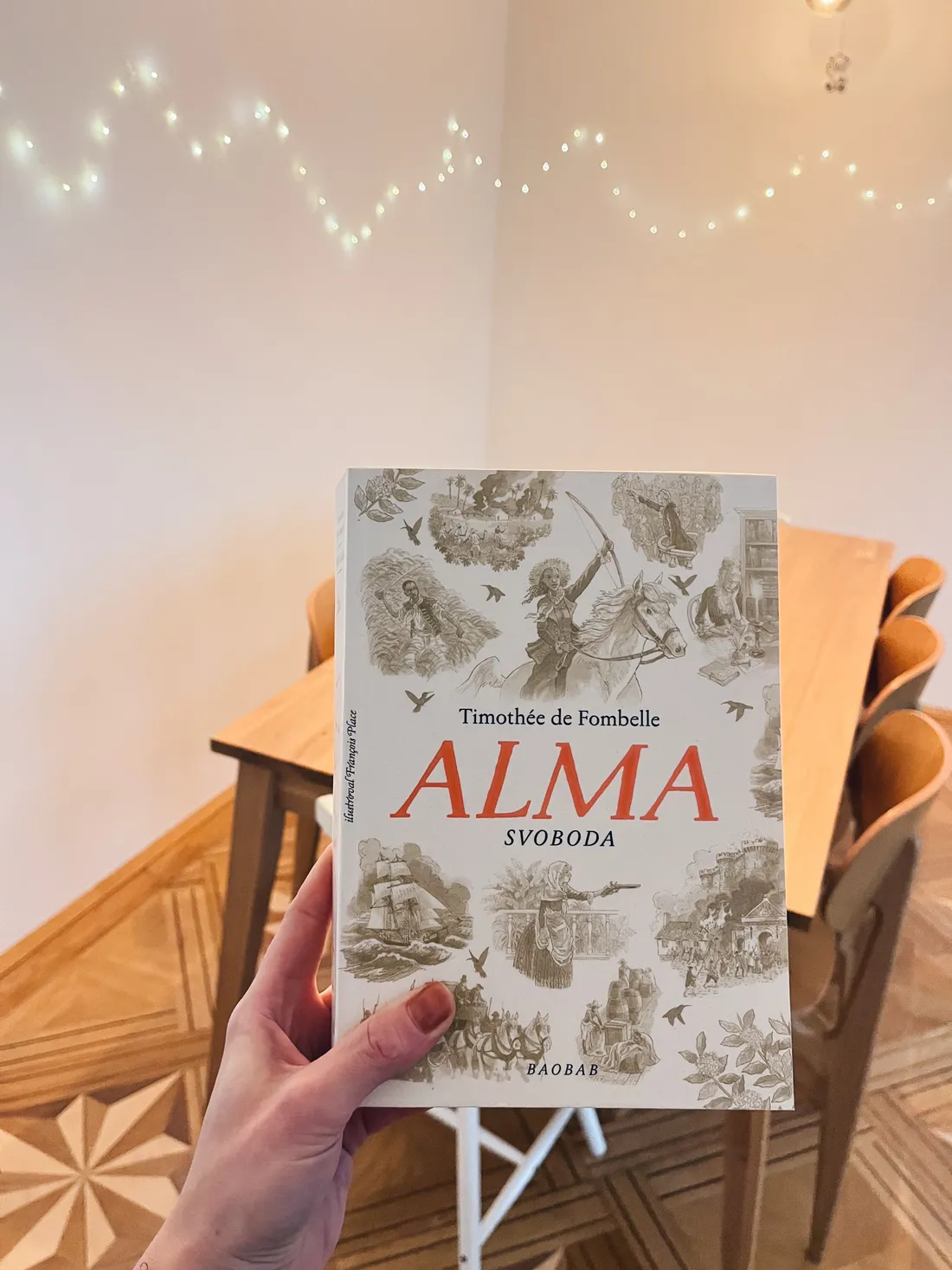 Ruka drží knihu Alma: Svoboda od Timothée de Fombelle s ilustracemi na bílé obálce v obývacím pokoji.