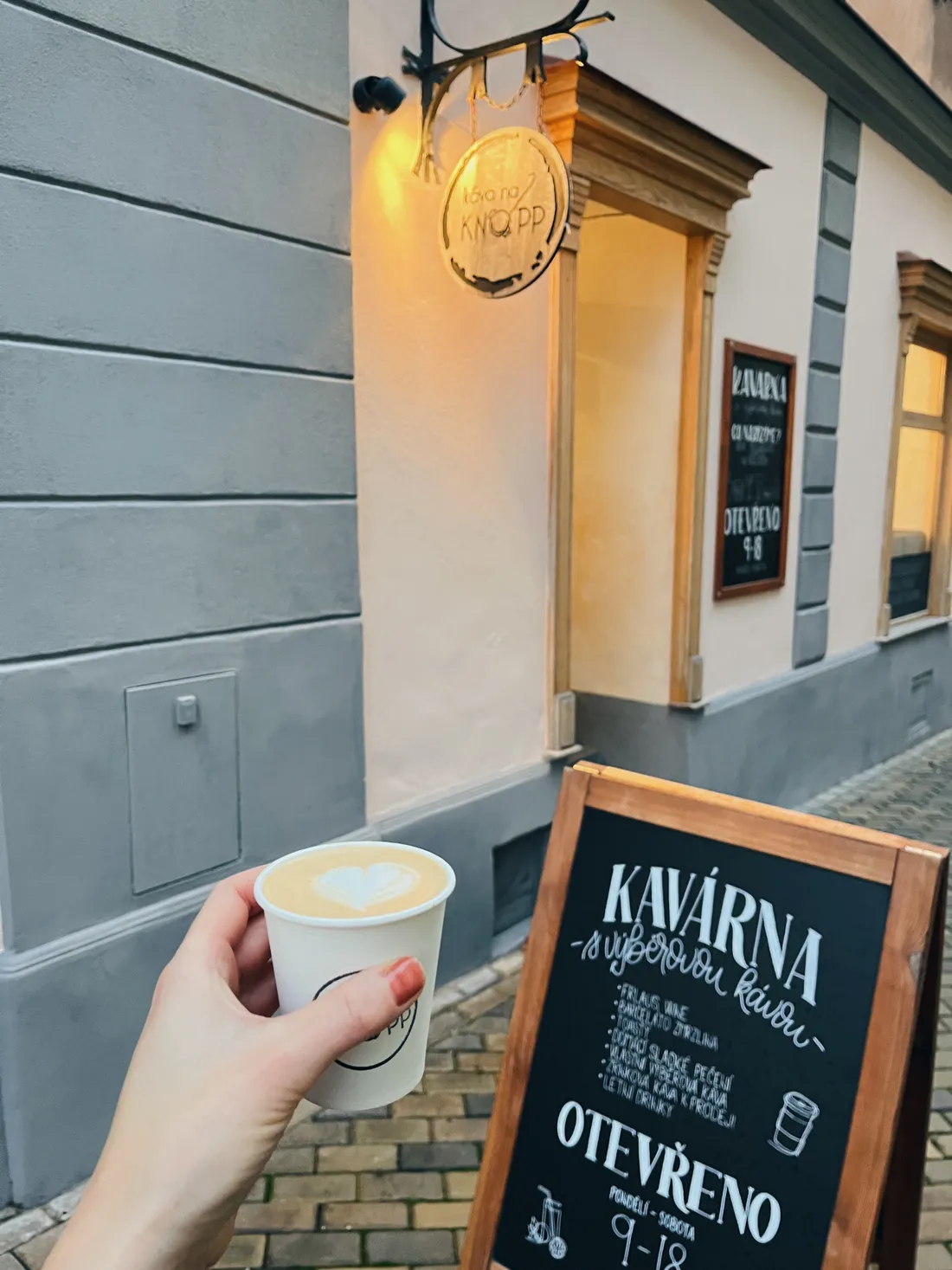 Bílý papírový kelímek s latte art srdcem před vchodem kavárny Káva na Knopp na dlažbě.