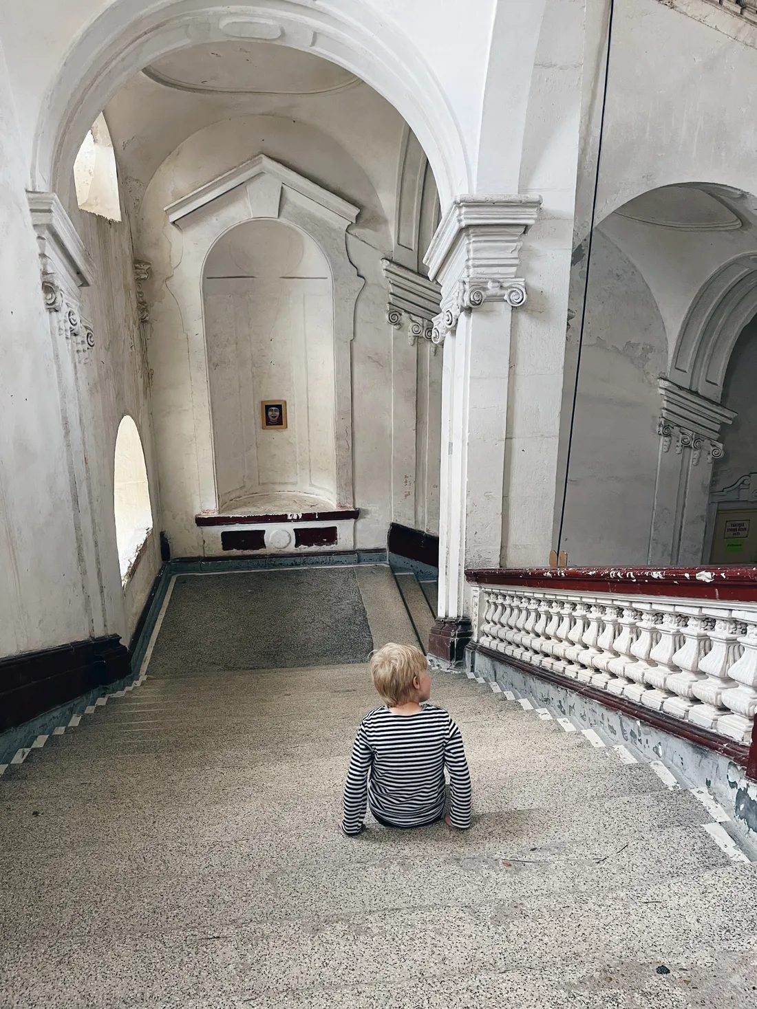 Blond dítě v pruhovaném tričku sedí zády na mramorovém schodišti starého paláce.