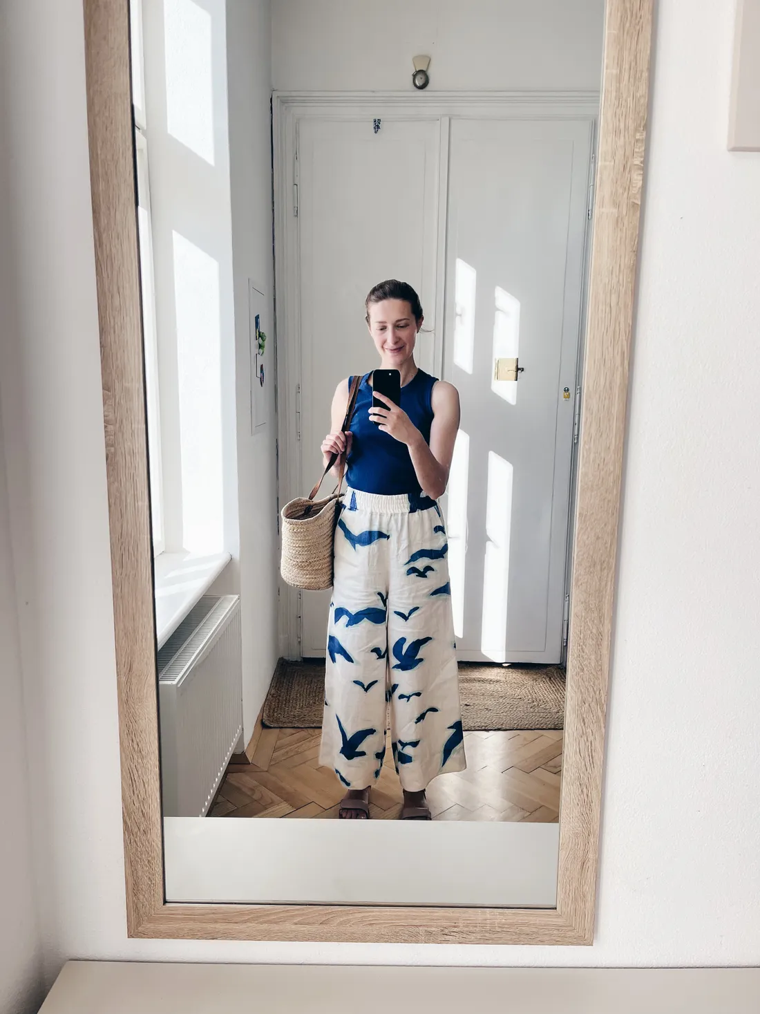 Selfie ve zrcadle: žena v tmavě modrém tílku, bílých kalhotách s modrými ptáky, proutěné tašce a parketové podlaze.