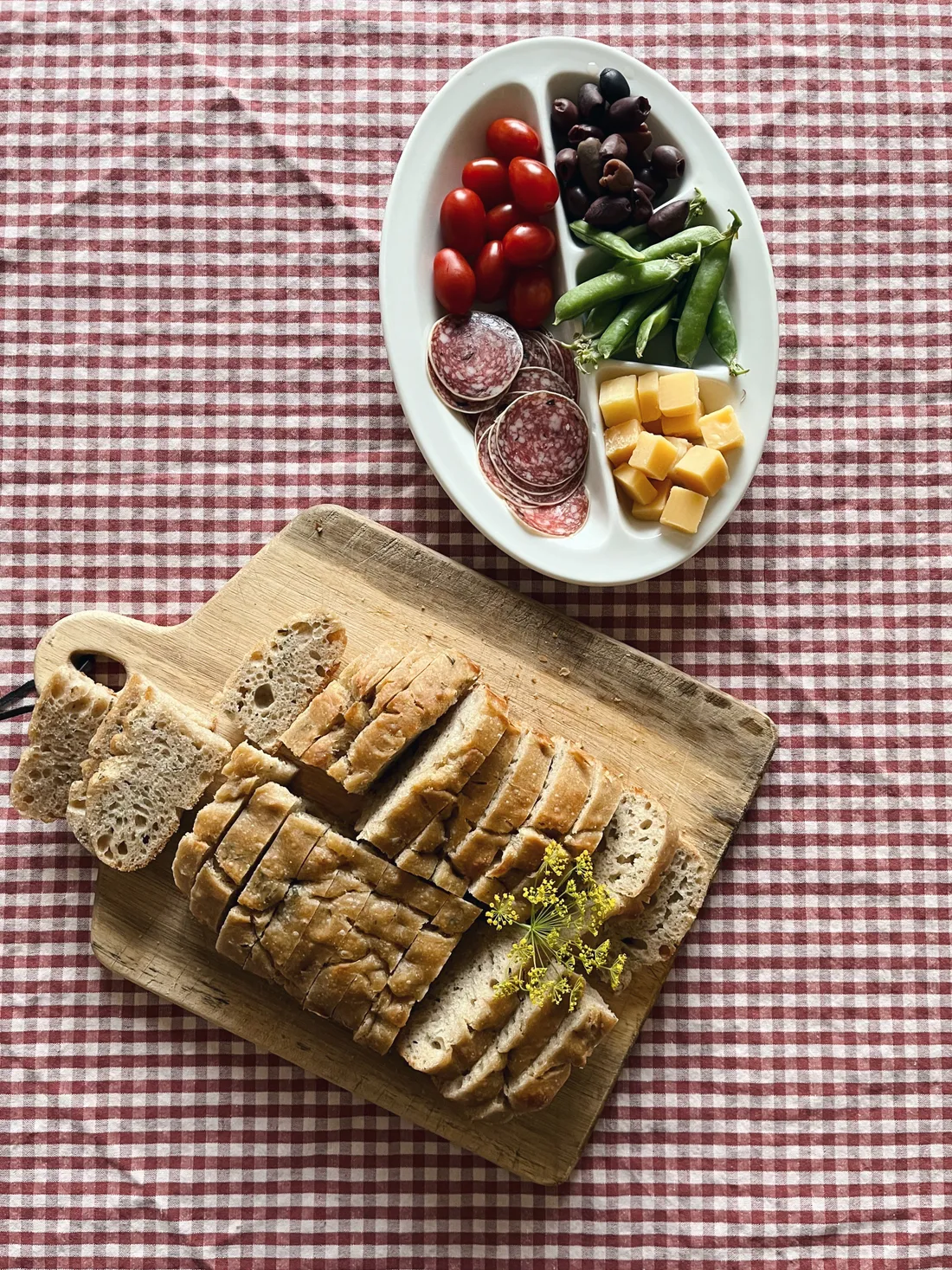 Plátky kváskového chleba na prkénku a mísa s rajčaty, olivami, sýrem a salámem na kostkovaném ubrusu.