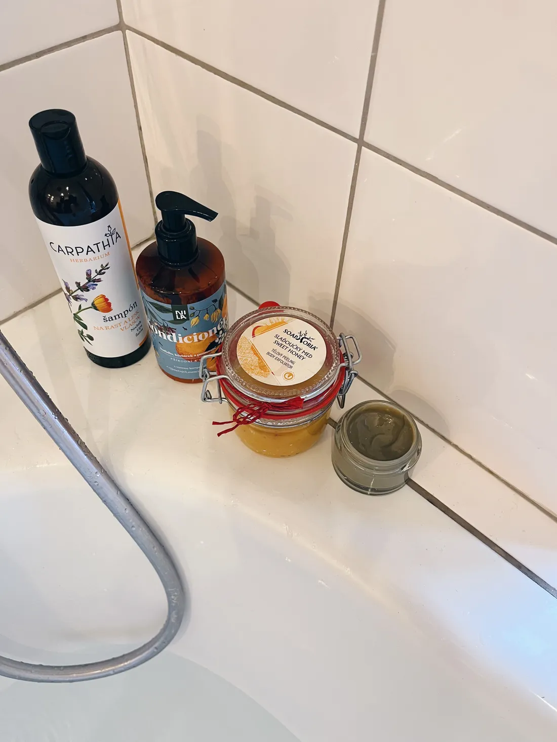Čtyři kosmetické produkty na bílém okraji vany — šampon, kondicionér, peeling a sklenička s maskou.