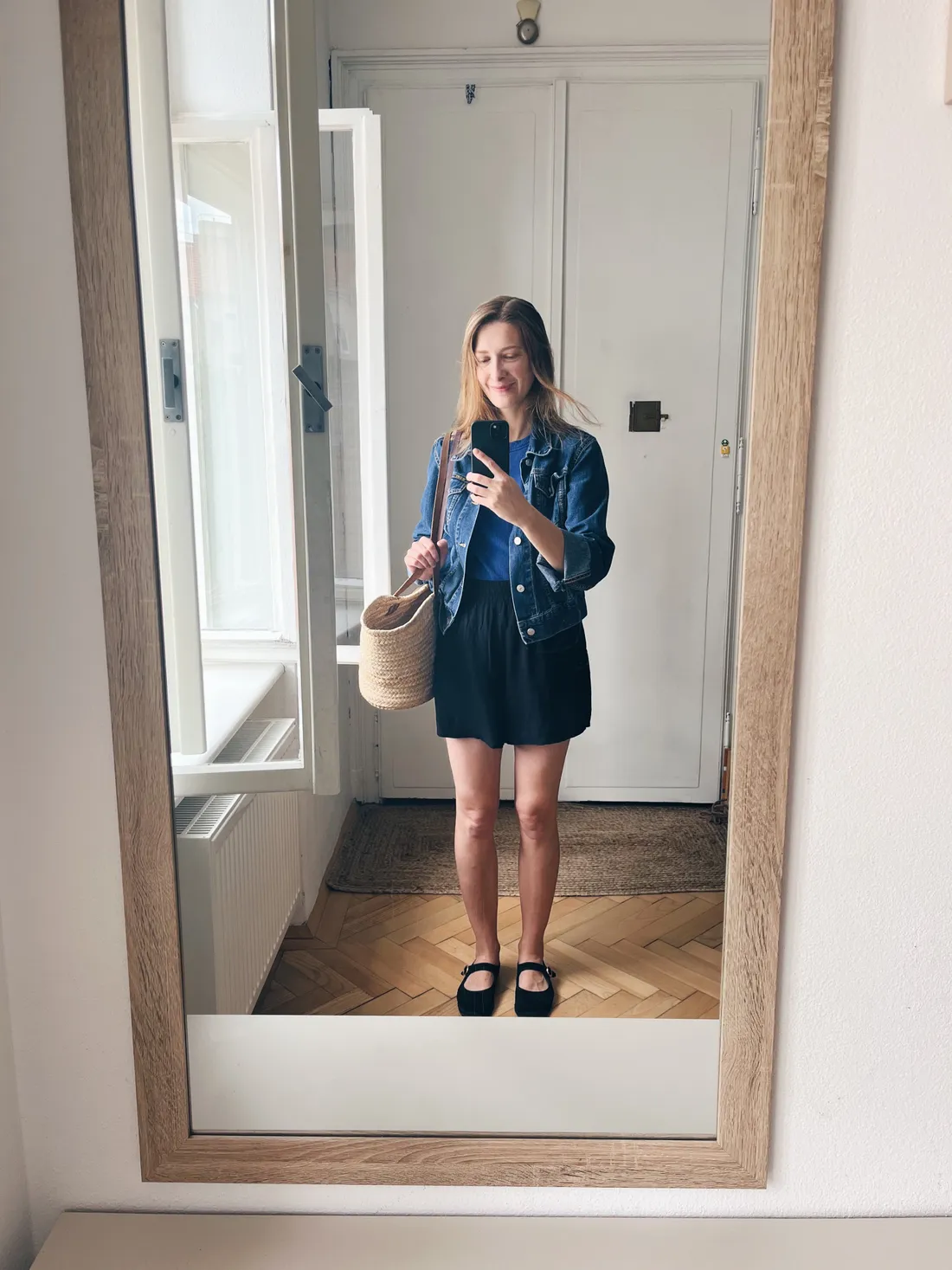Žena v džínové bundě a černých šortkách se slaměnou taškou v zrcadlové selfie v předsíni.