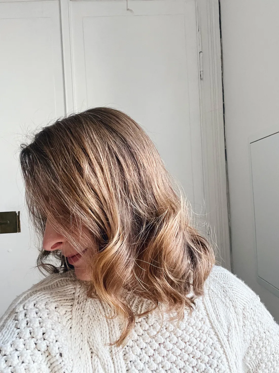 Detail vlnitých blond vlasů a světlého pleteného svetru, žena se dívá dolů k bílým dveřím
