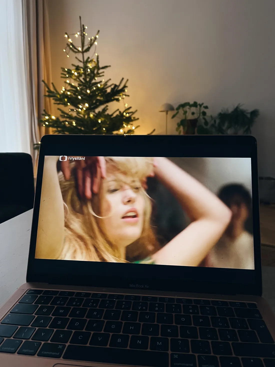 MacBook Air s českou klávesnicí přehrává iVysílání, za laptopem vánoční stromek se žlutými světýlky