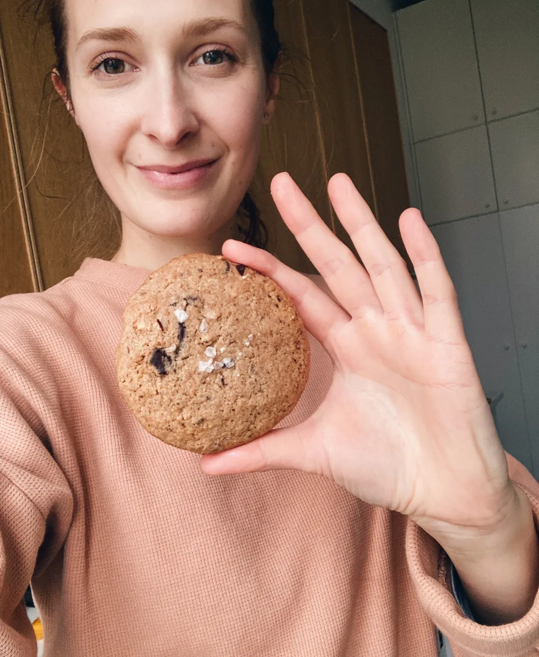Autorka drží v ruce kulatou lískooříškovou cookie s kousky čokolády a vločkami maldonské soli, usmívá se do objektivu.