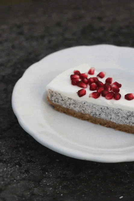 Kus makového cheesecaku s bílým krémem posypaný granátovým jablíčkem na bílém talíři.