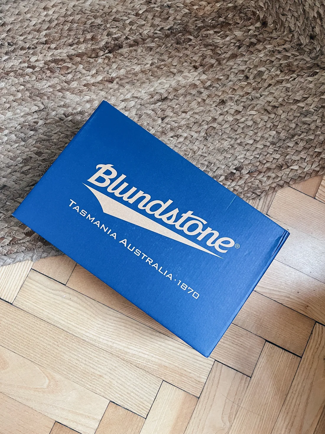 Modrá krabice od bot Blundstone s bílým logem na koberci a parketách.
